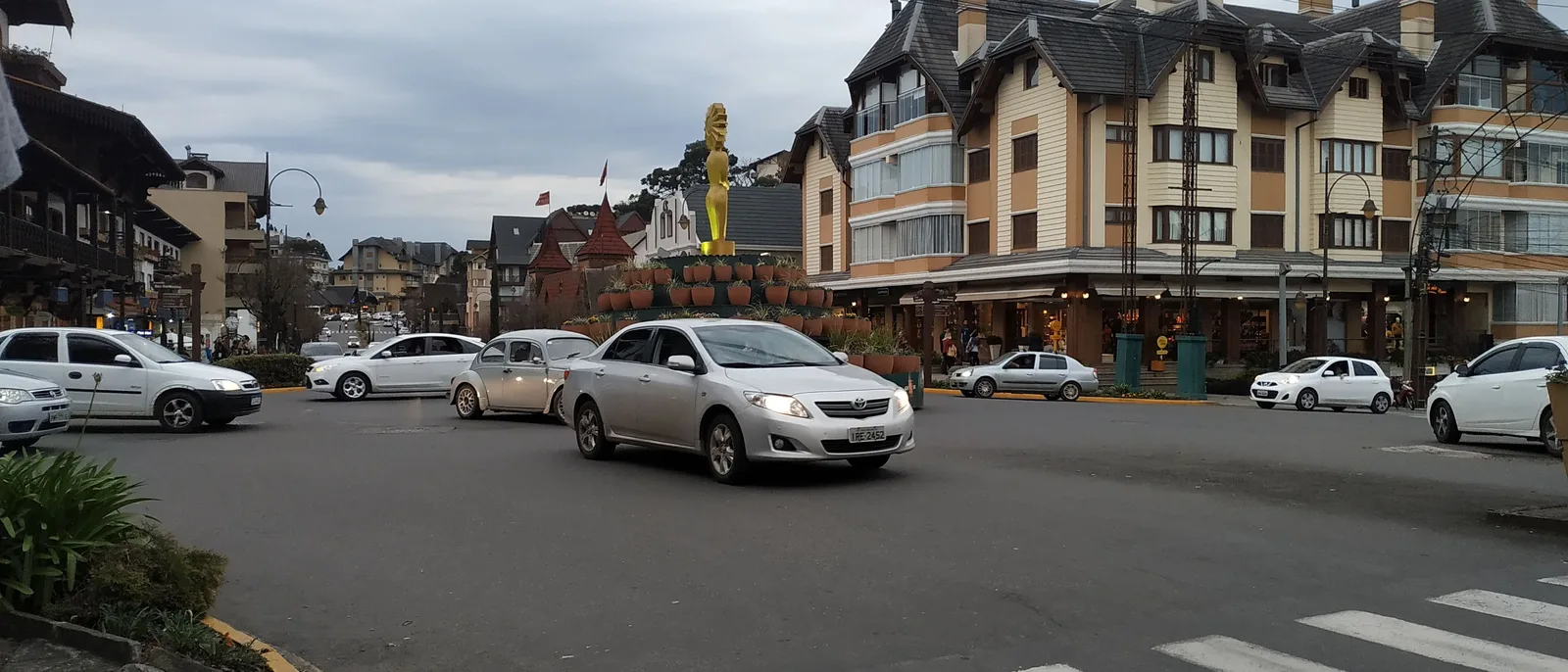 A corner of the Avenida Borges de Medeiros in central Gramado on a gray day: cars at an intersection in front of a tall half-timbered apartment building and a golden statue on a flowerbed
