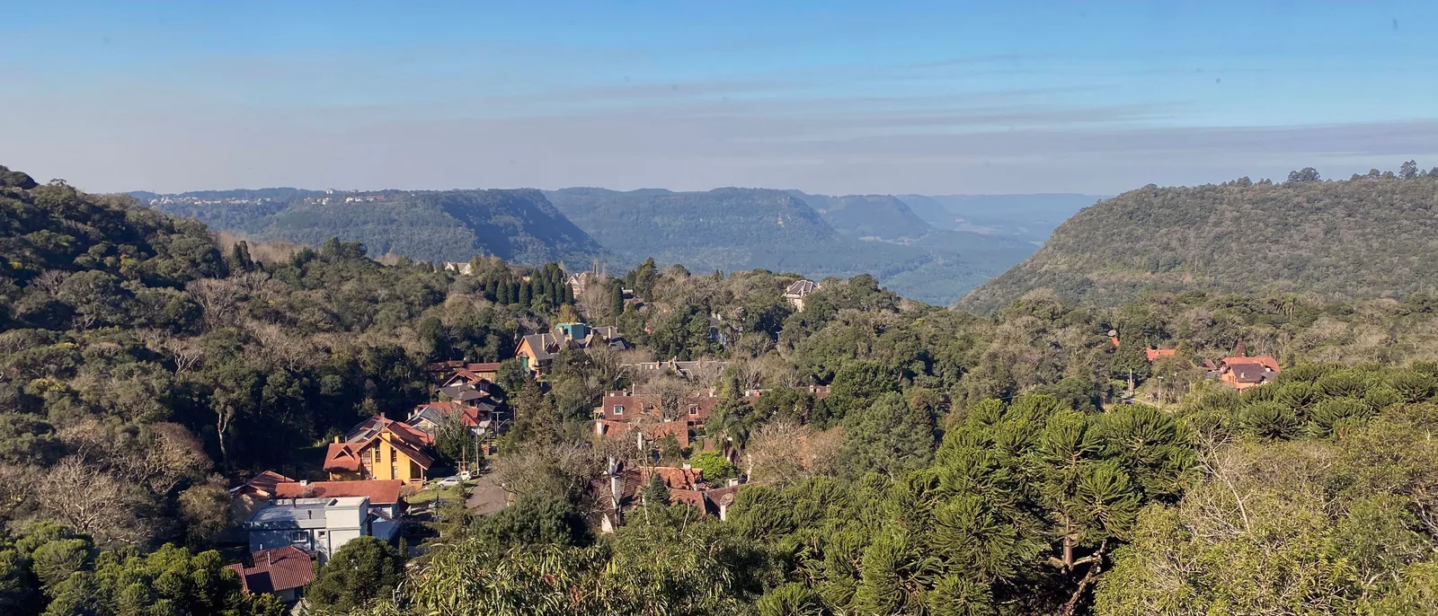 Houses with steep red roofs among dense trees in Gramado’s Planalto district, with the long ridges of the Vale do Quilombo fading into haze behind