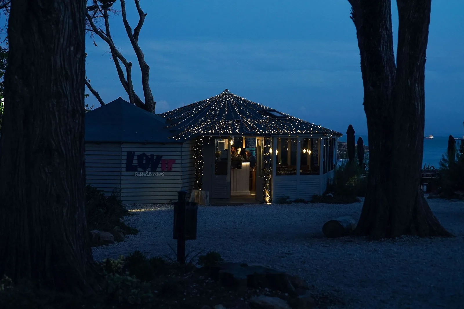 A seaside restaurant strung with lights after dark, tables set out under them