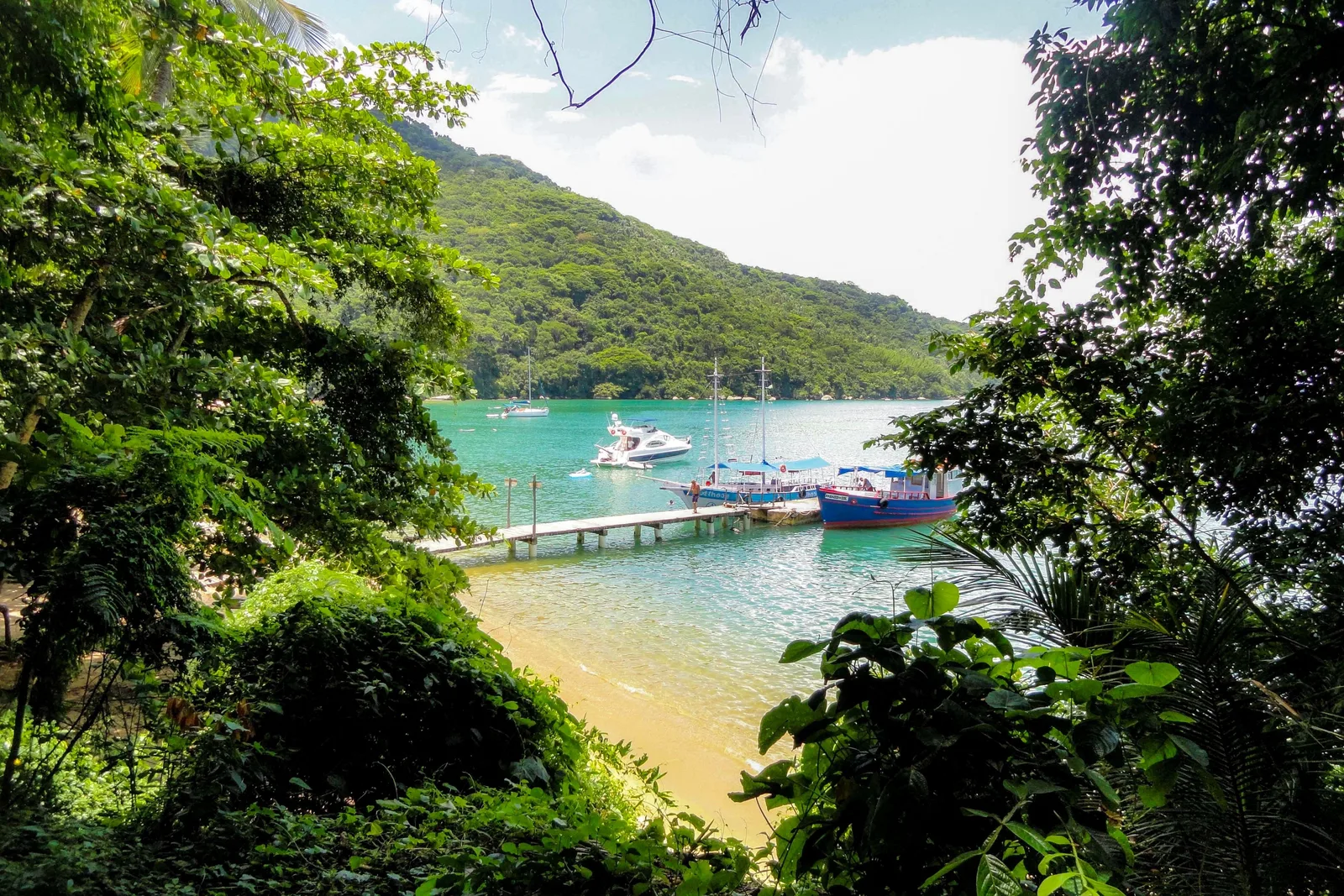 A jetty on Ilha Grande seen through the trees, boats moored off a small beach in clear turquoise water