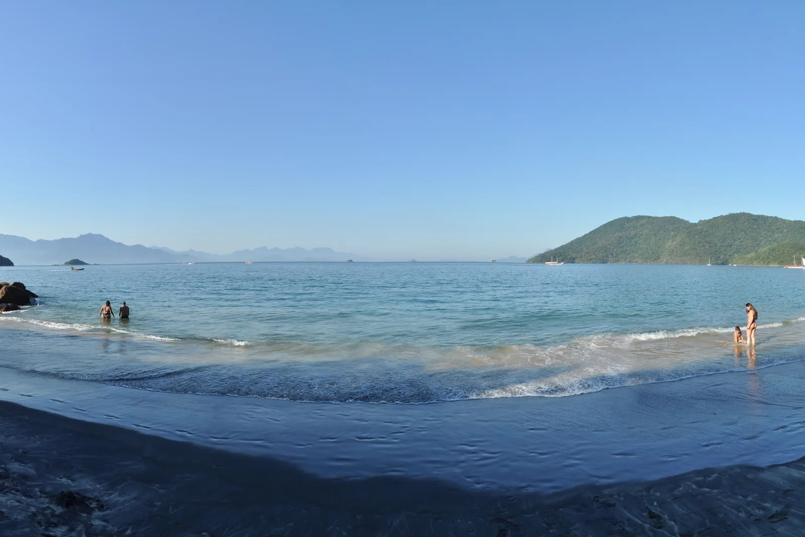 A dark-sand beach on Ilha Grande with shallow water running up it and forested headlands closing the bay