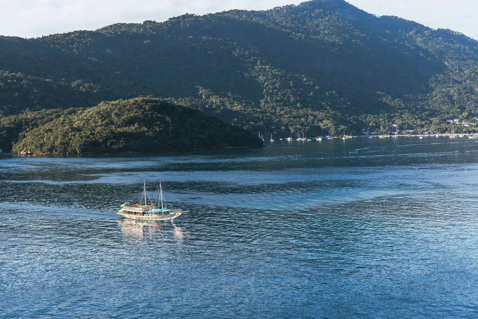 A boat under sail on flat blue water with the island’s forested hills behind