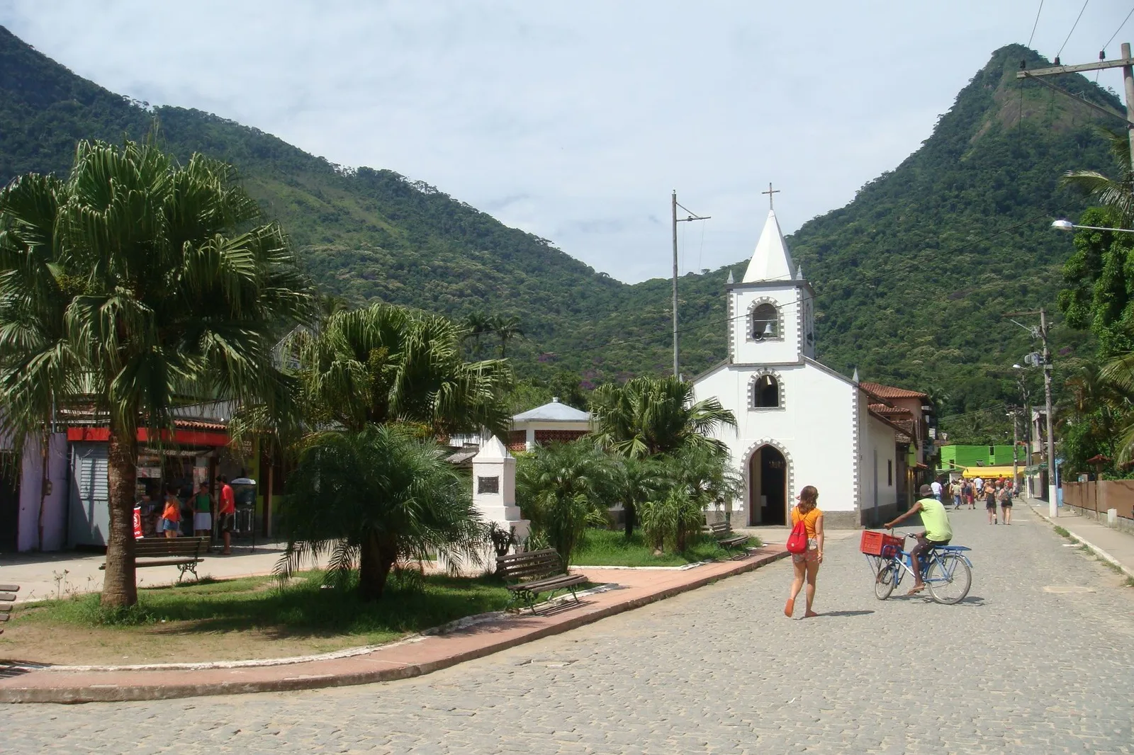 The white church with its pointed steeple on the main square of Vila do Abraão, palms and a paved street in front and forested peaks behind