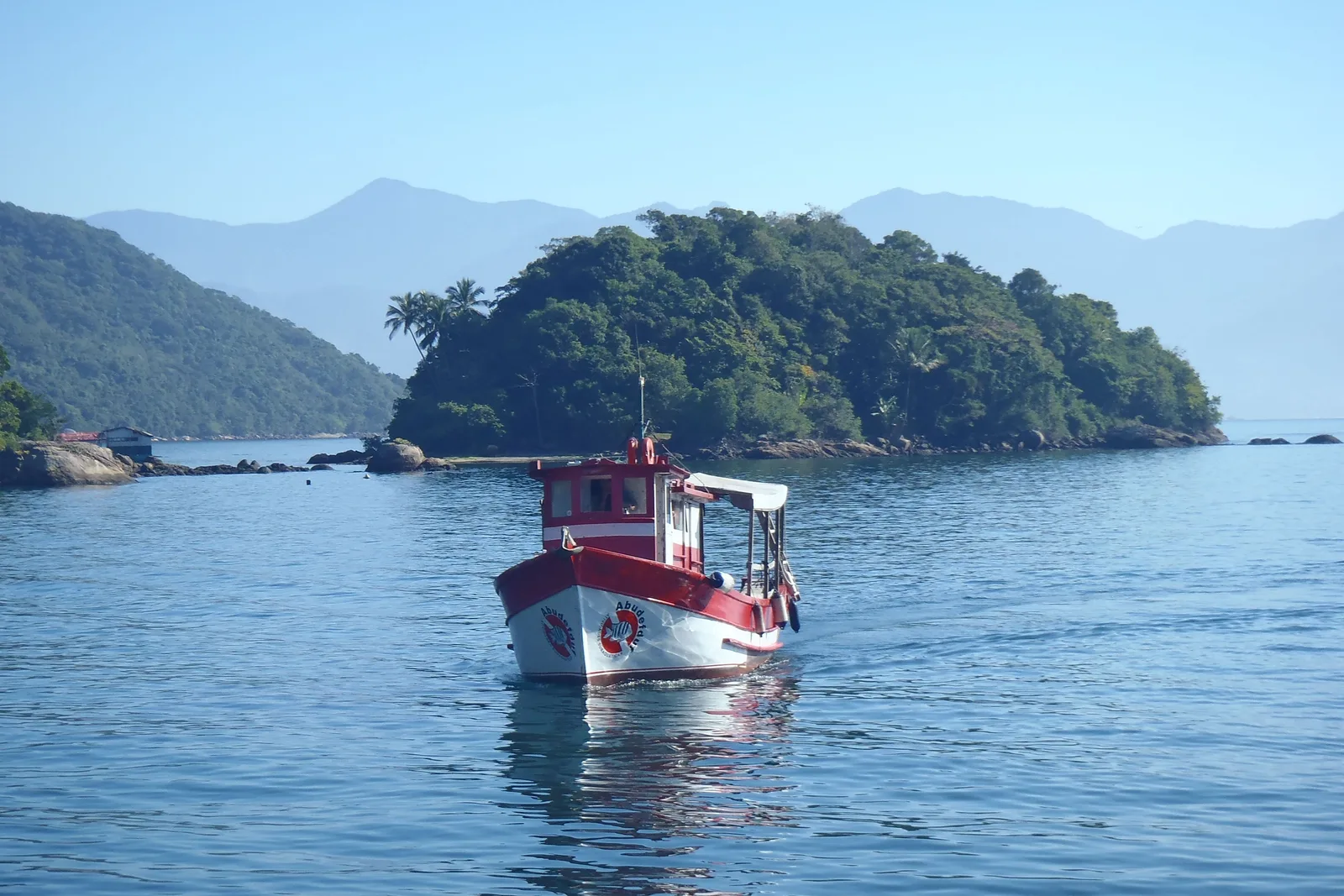 A red and white fishing boat on still water in front of a small forested island