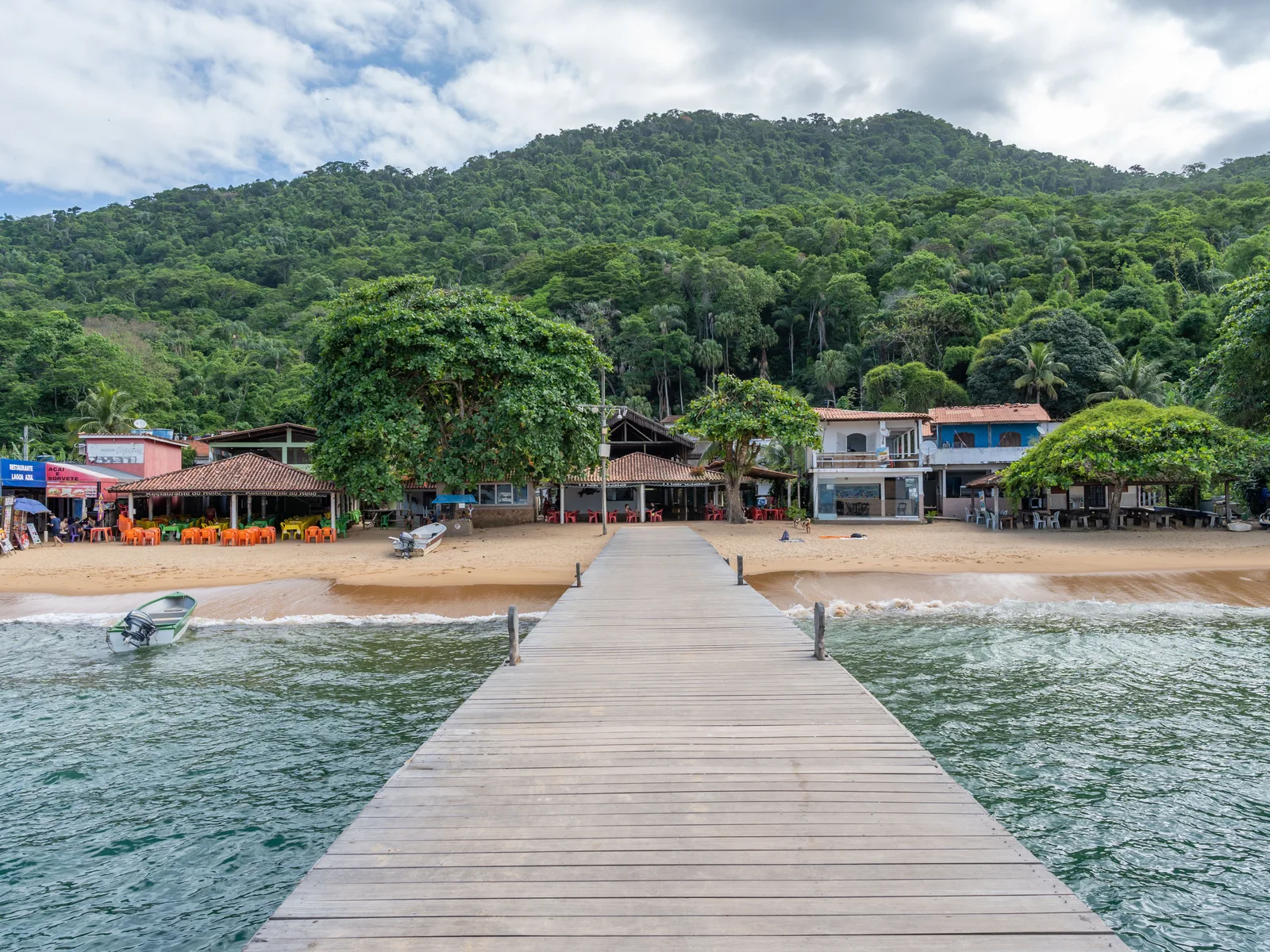 A wooden jetty running out to a beach on Ilha Grande, low buildings and forested hills behind it