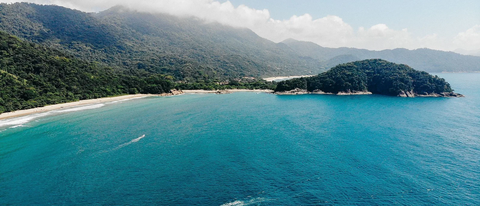 A wide beach on the island’s ocean side seen from the air, forested headlands closing it at both ends