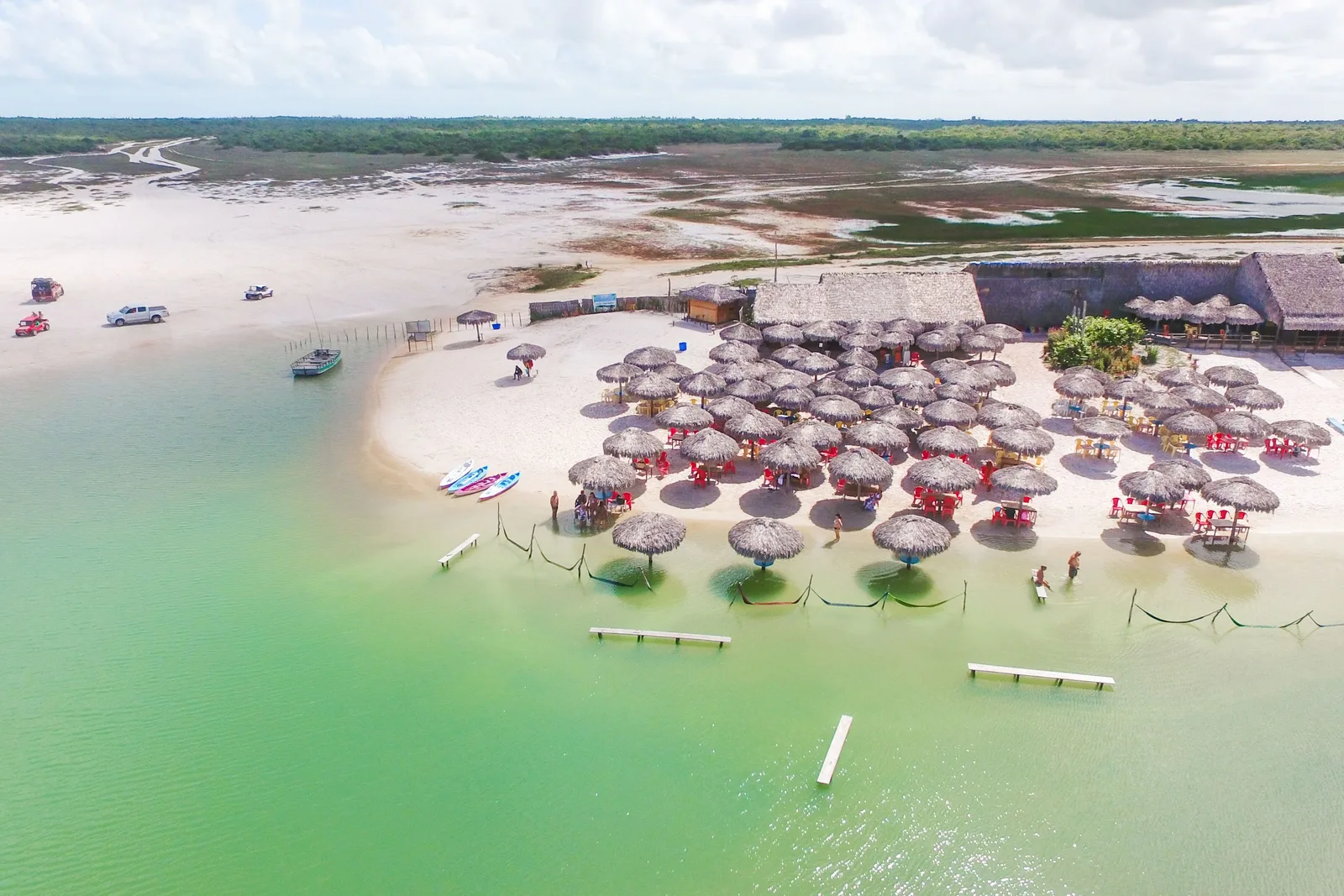 Rows of thatched parasols and a wooden deck set out along the shallow green edge of Lagoa Azul, seen from above, with a sandbank curving into the water