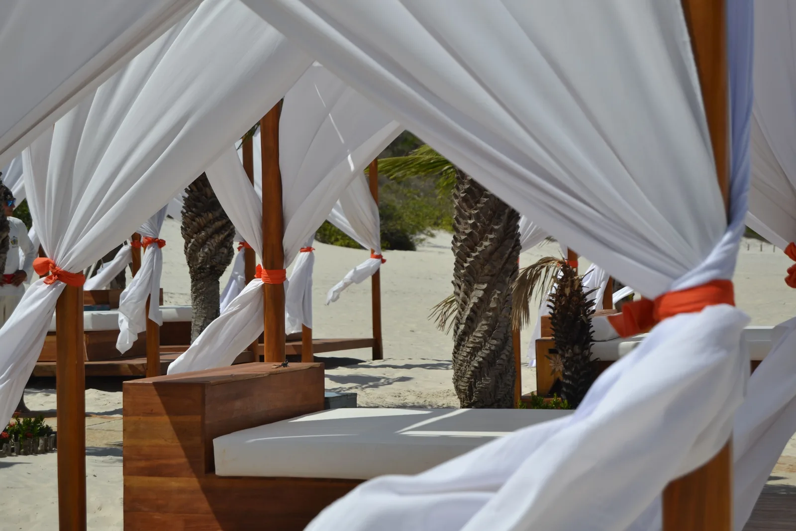 A row of wooden four-poster daybeds with white curtains tied back, set out on the sand at the edge of the lagoon under palm trunks