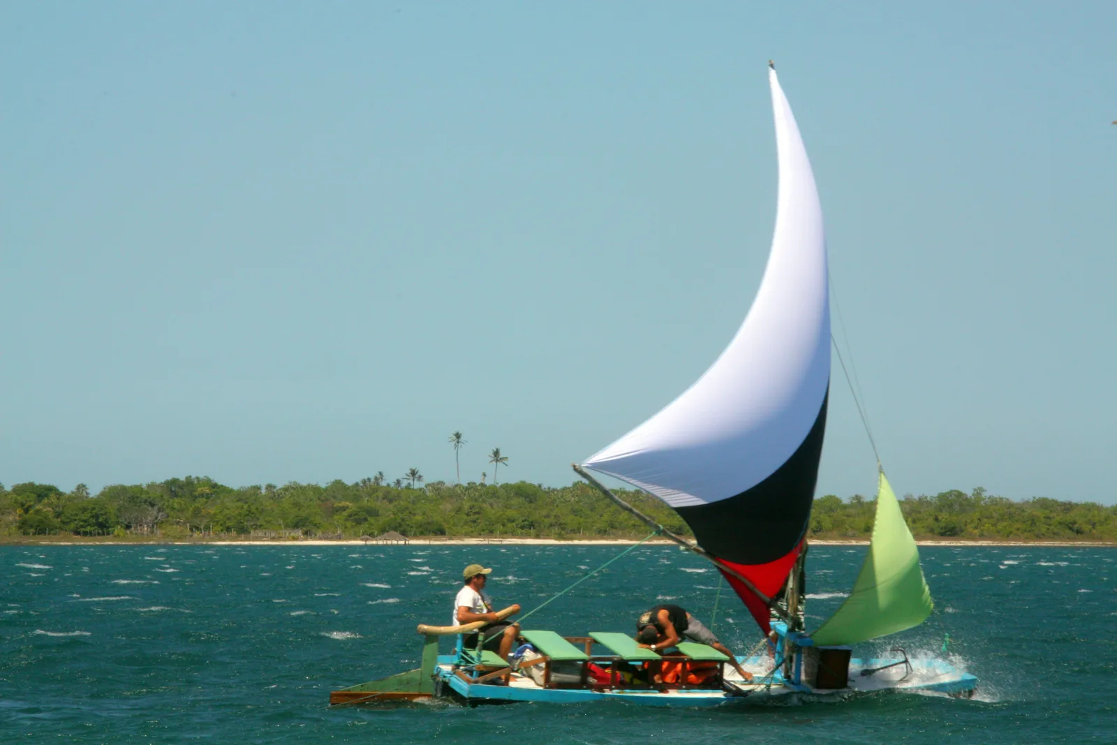 A wooden jangada with a tall white and dark sail crossing the flat blue water of the lagoon, two people aboard and a low green shore behind