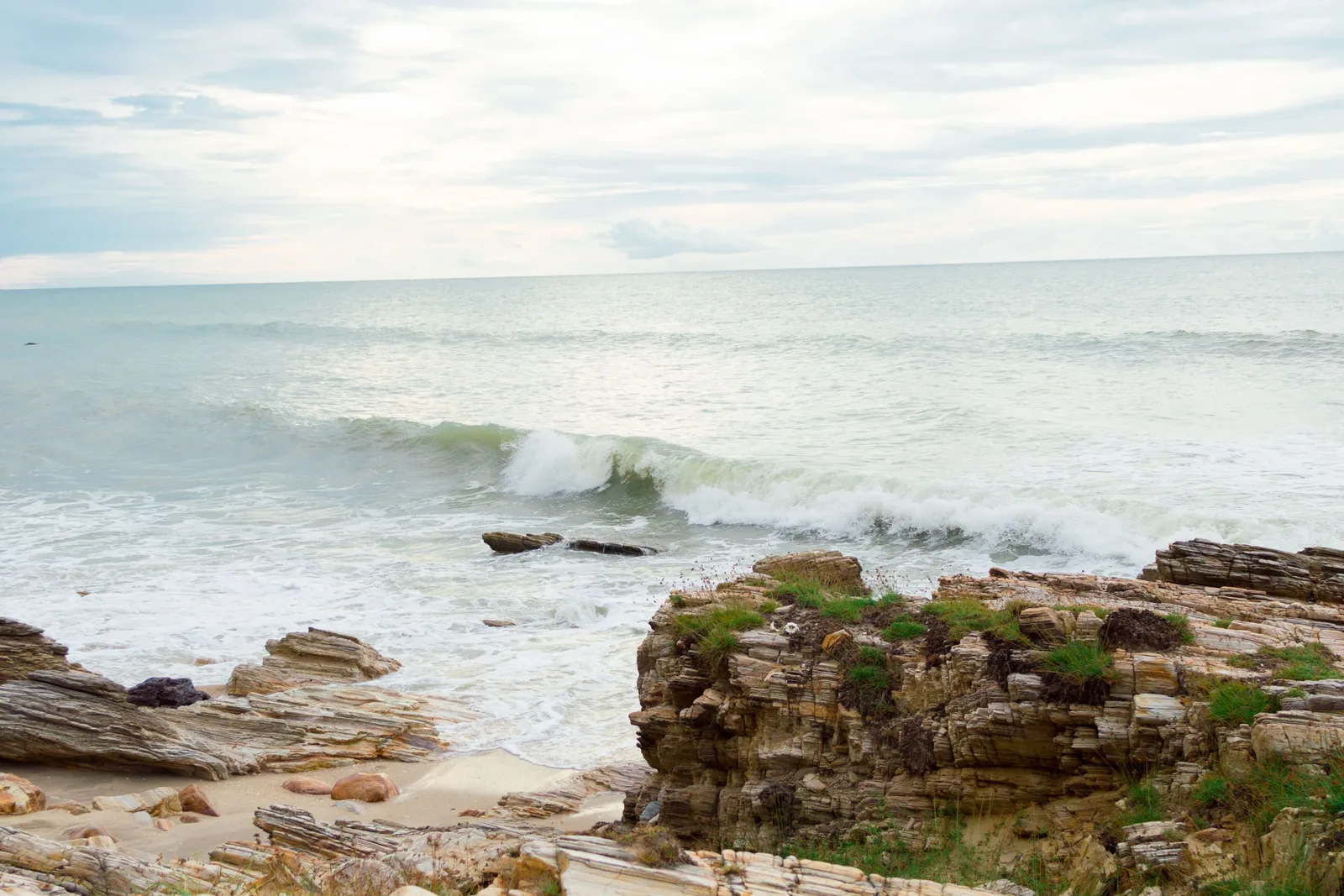 Layered dark rock shelves running into the sea at Praia da Malhada with white surf breaking over them under an overcast sky
