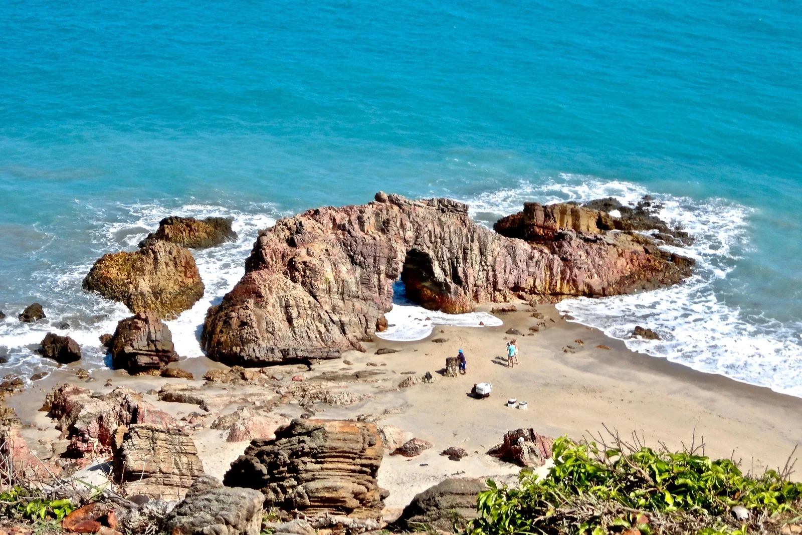 The rocky headland at Jericoacoara seen from above, dark banded rock and the arch of Pedra Furada standing in white surf with turquoise water beyond and green scrub in the foreground