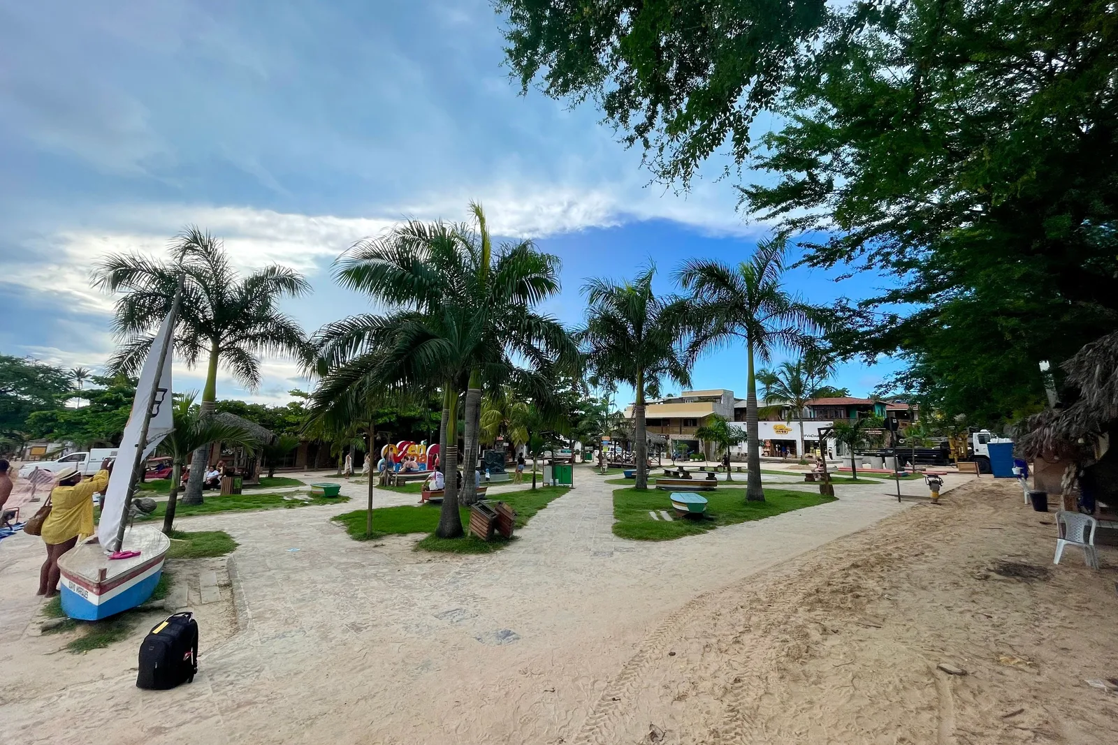 An open sand square at Jericoacoara with a small boat and a mast on it, tall coconut palms, low white buildings behind and a bright sky with scattered cloud