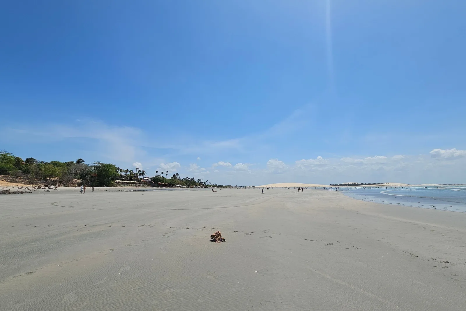 A wide open beach at Jericoacoara under a hazy sky, pale sand running out to a flat sea with a low green shoreline in the distance