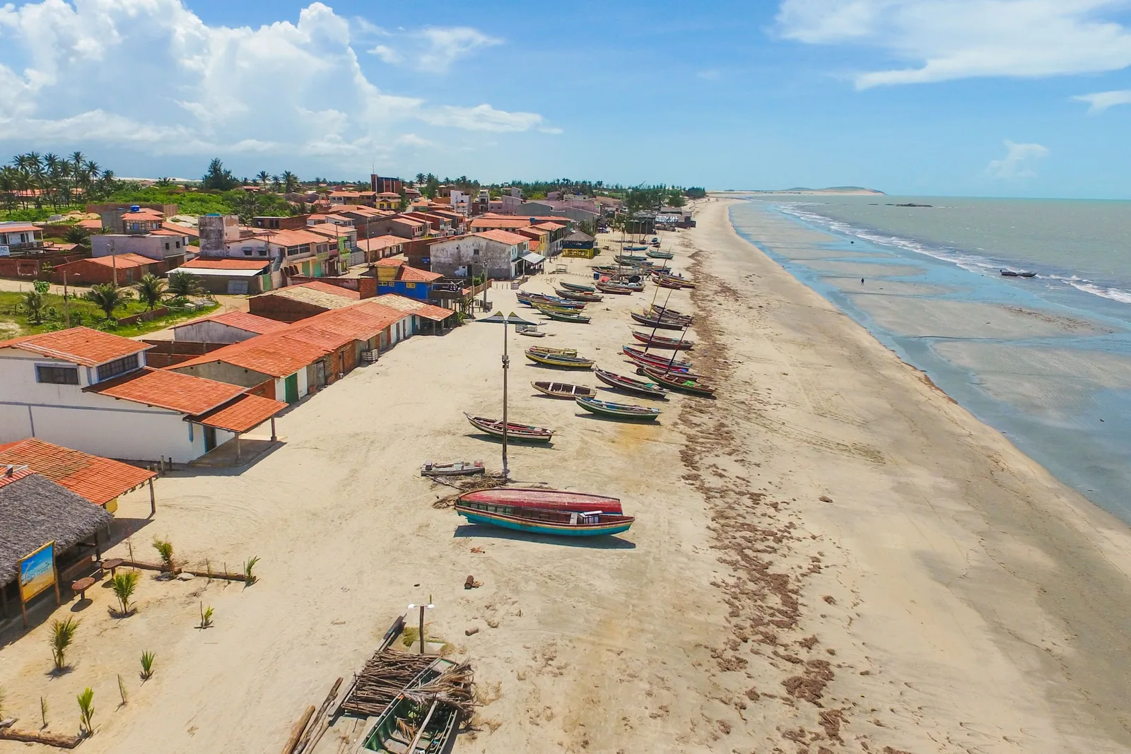 The village of Praia do Preá from the air: red-roofed houses and wooden fishing boats along the top of a long pale beach running away into the distance beside a flat sea