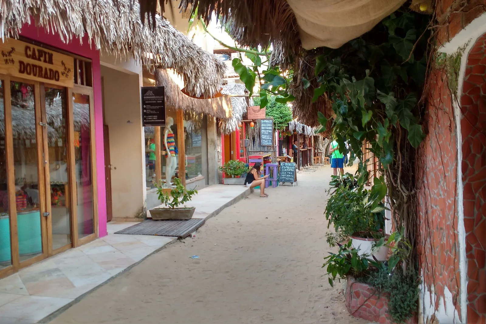 A sand street in Jericoacoara with pink, turquoise and cream shopfronts along one side under a thatched awning, a stone-paved walkway in front of them and people in the distance