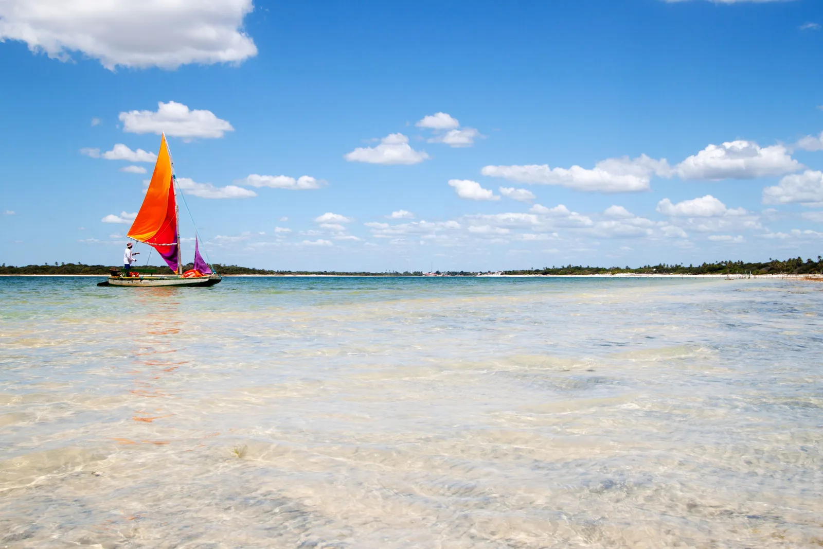 A jangada with a bright orange and red sail crossing the shallow pale-green water of the Tatajuba lagoon under a blue sky with scattered cloud