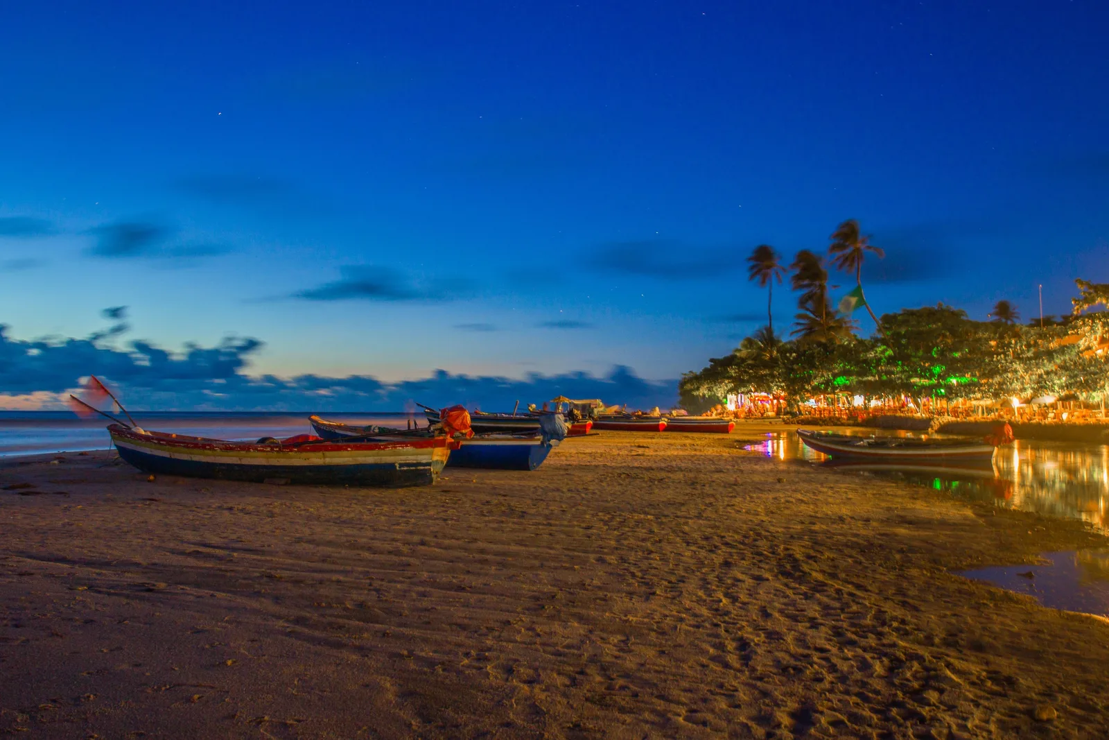 Fishing boats drawn up on dark sand at dusk with the warm lights of the village strung along behind them under a deep blue sky