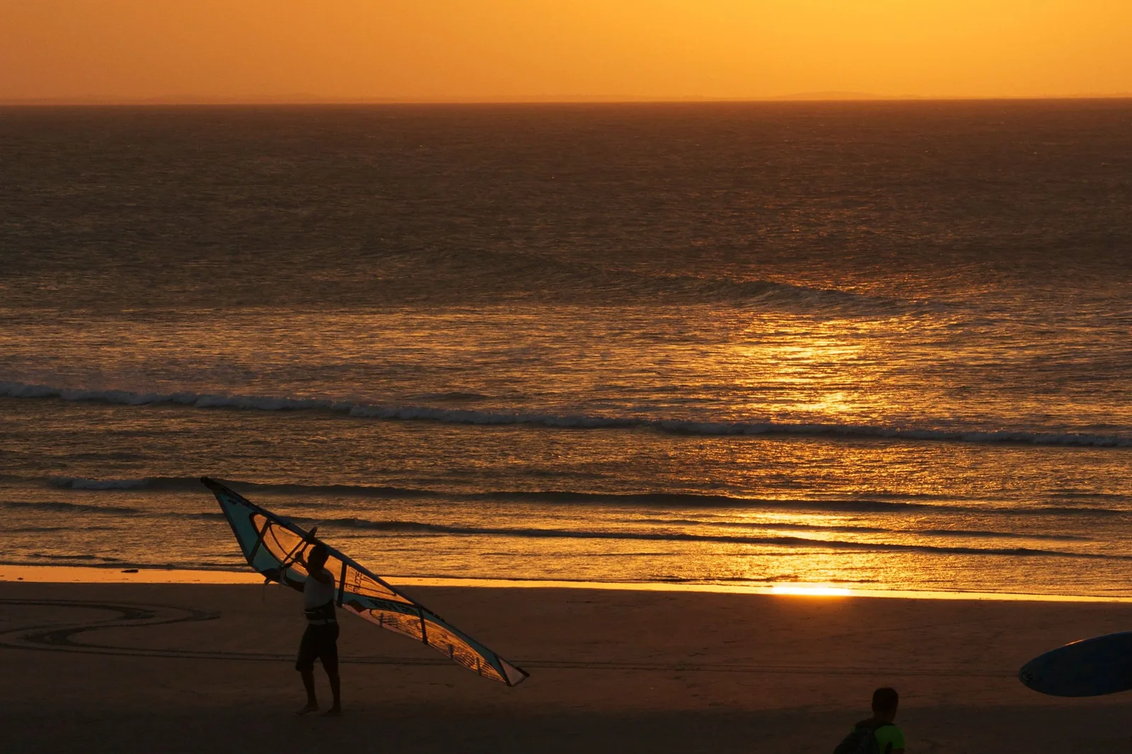 A windsurfer carrying a rig along the beach in silhouette against a low sun, the sea flat and gold behind them