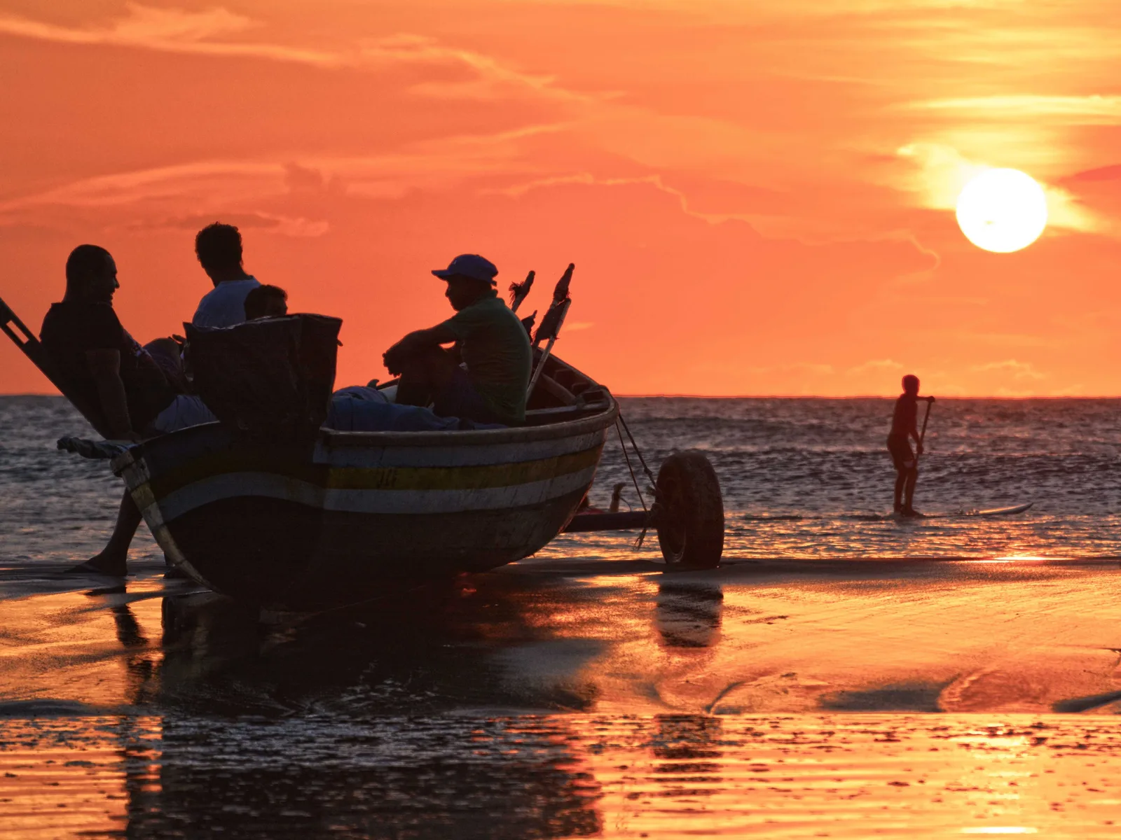 People sitting in a wooden boat drawn up on the sand watching an orange sunset over the sea, with one figure standing at the water’s edge
