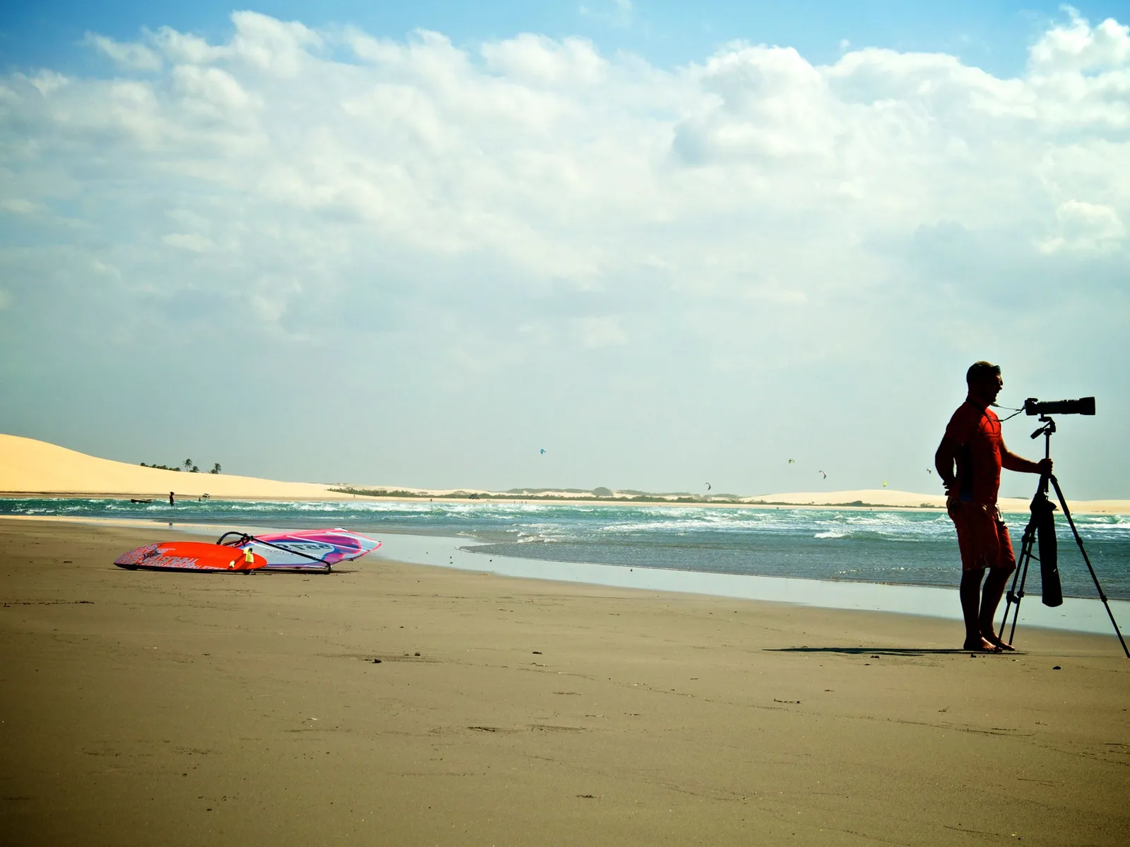 A photographer standing at a tripod on the beach in the late sun with a windsurf board and its sail rig laid out on the sand beside them, and kites out over the water beyond