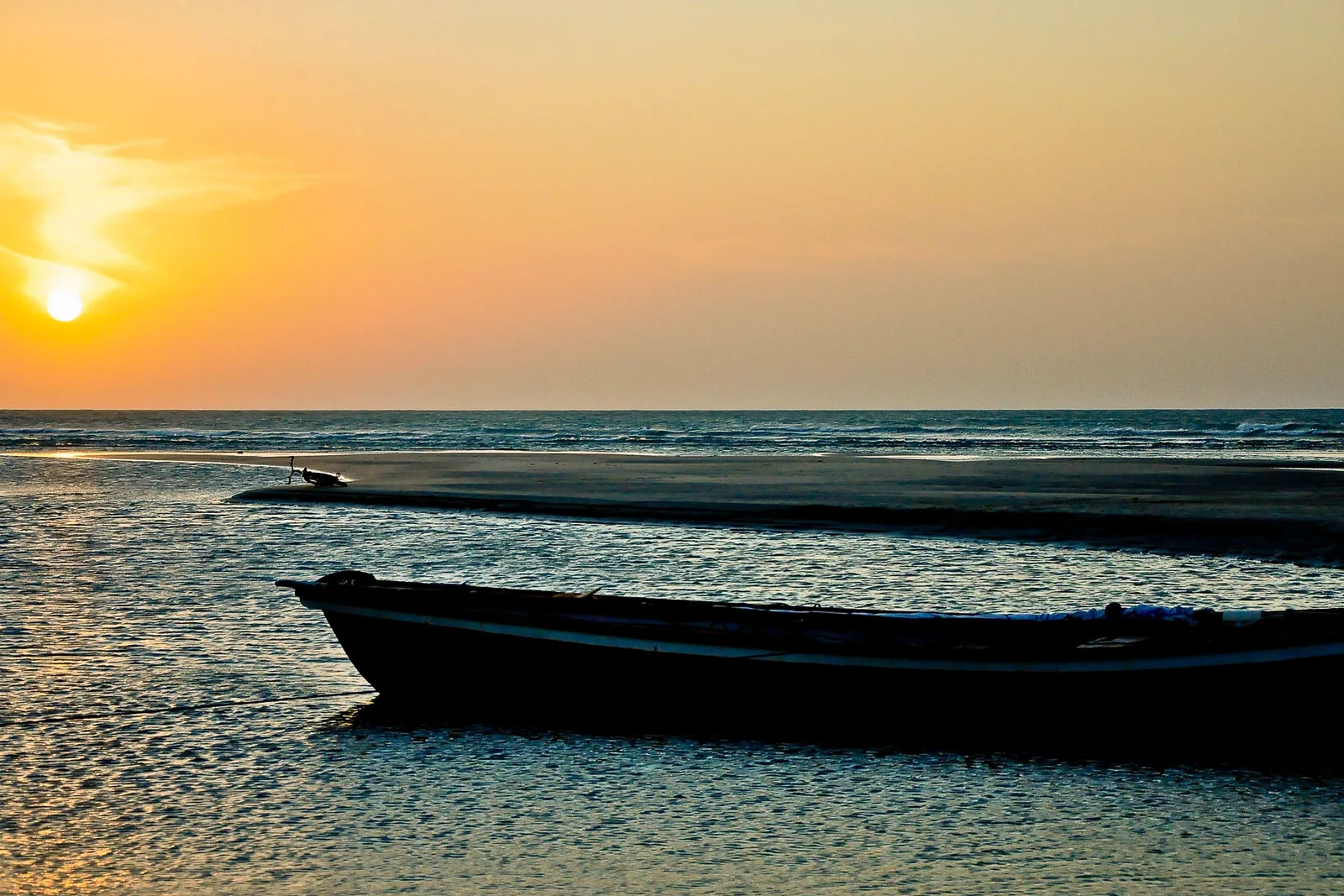 A wooden fishing boat drawn up on a wet sand flat at sunset, its hull dark against the reflected light