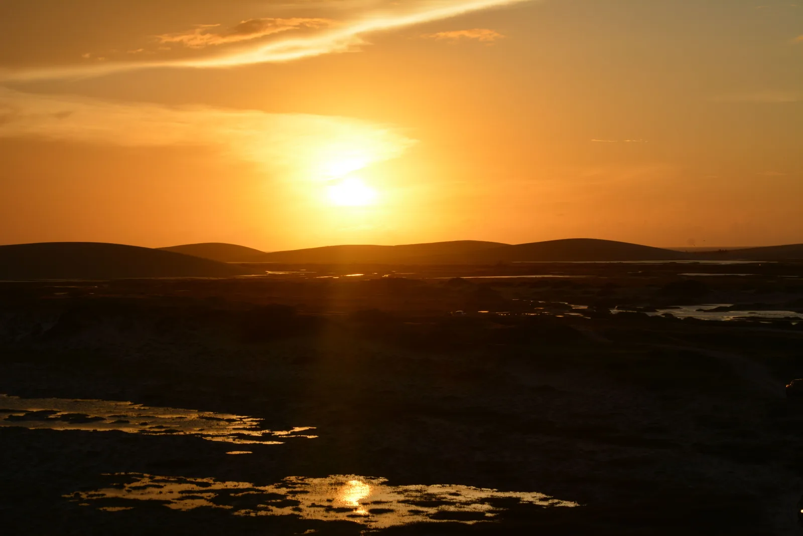 The sun setting behind a low dune ridge at Jericoacoara, the light spread flat across wet sand in the foreground