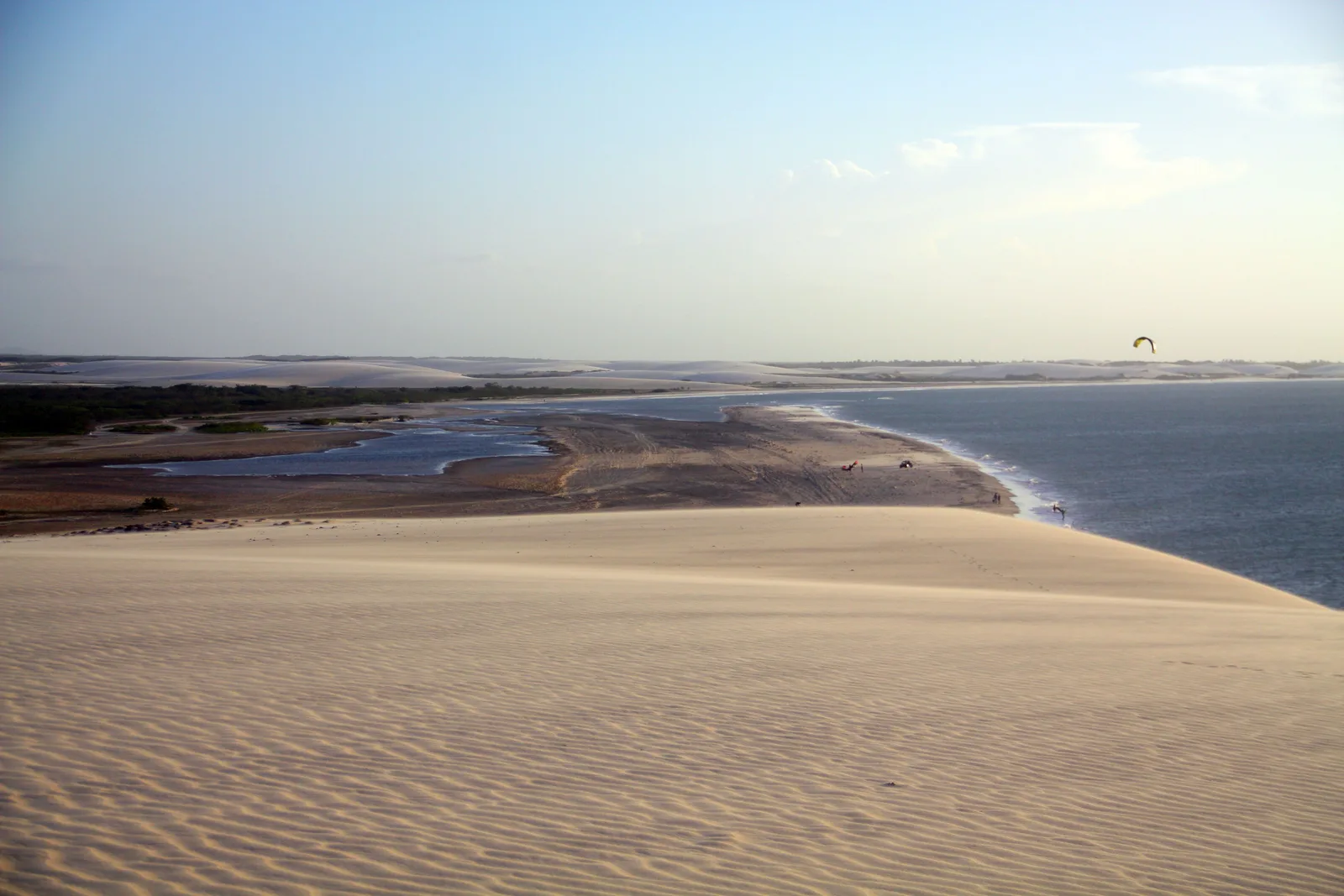 The slope of a pale dune running down to a strip of shallow water and the open sea, with one kite flying high above the far shore