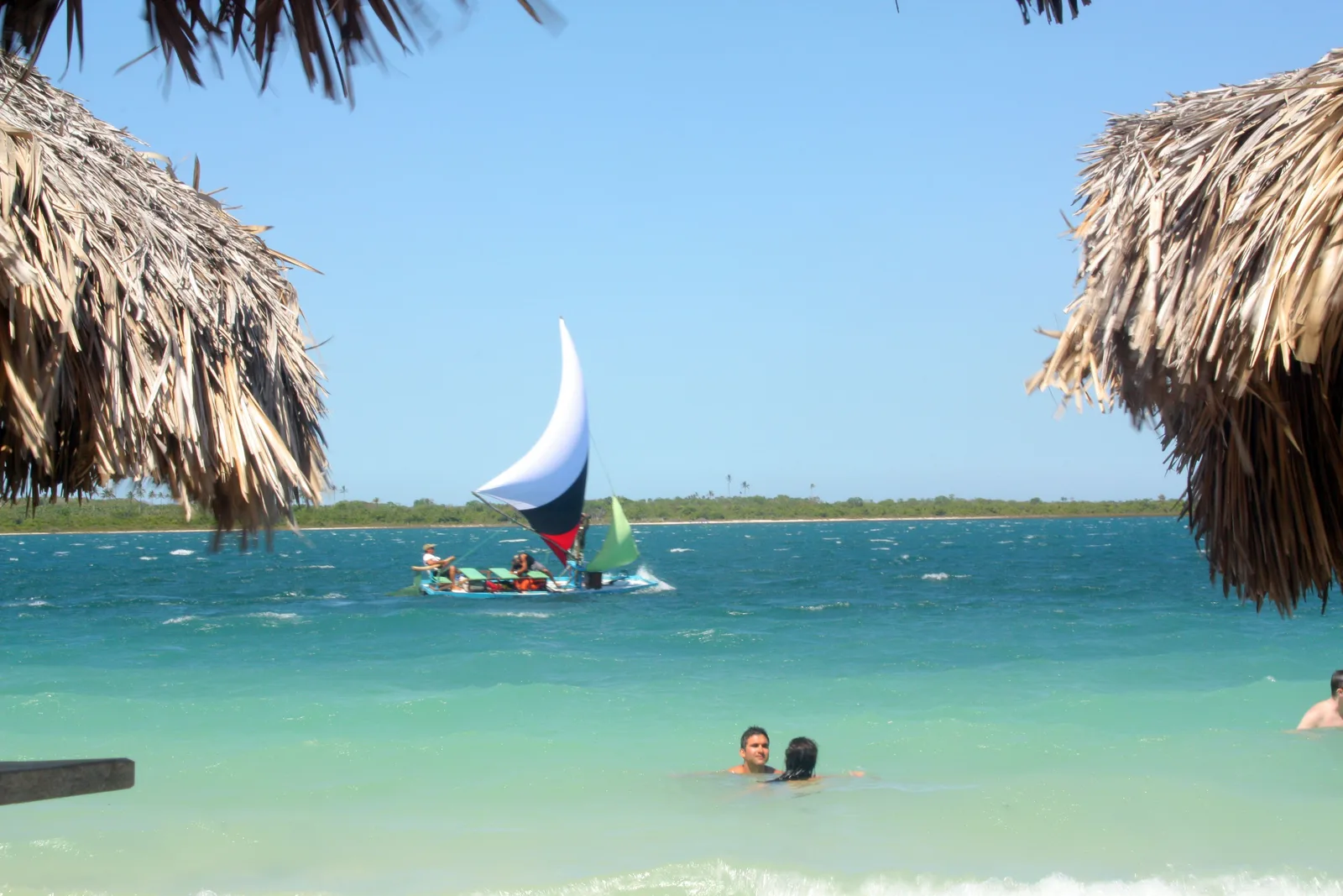 A small sailing boat on turquoise lagoon water seen between two thatched parasols, with swimmers standing in the shallows
