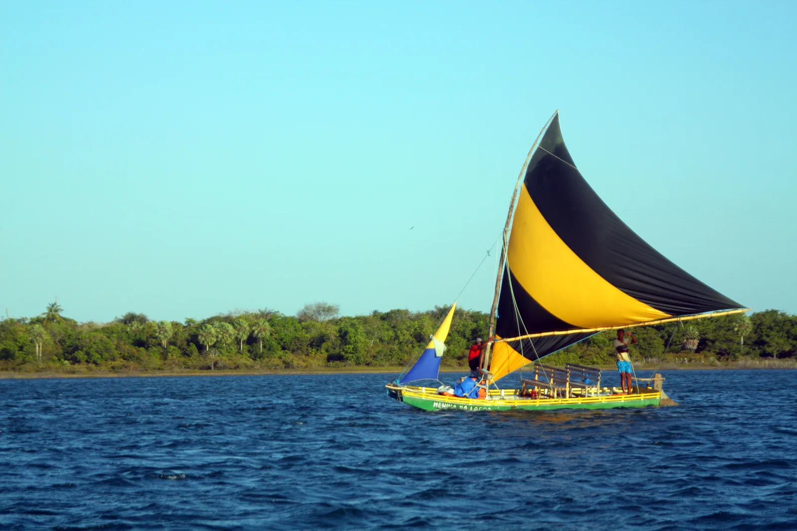A jangada with a yellow and black sail crossing deep blue lagoon water in front of a low green shore