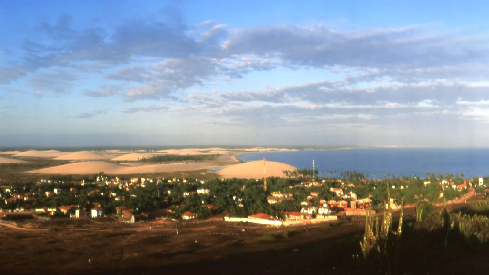 Jericoacoara from above in late afternoon sun: red roofs and green trees spread along the shore under a blue sky, a red and white communications mast standing among them, the dune field running away to the left and open blue sea to the right