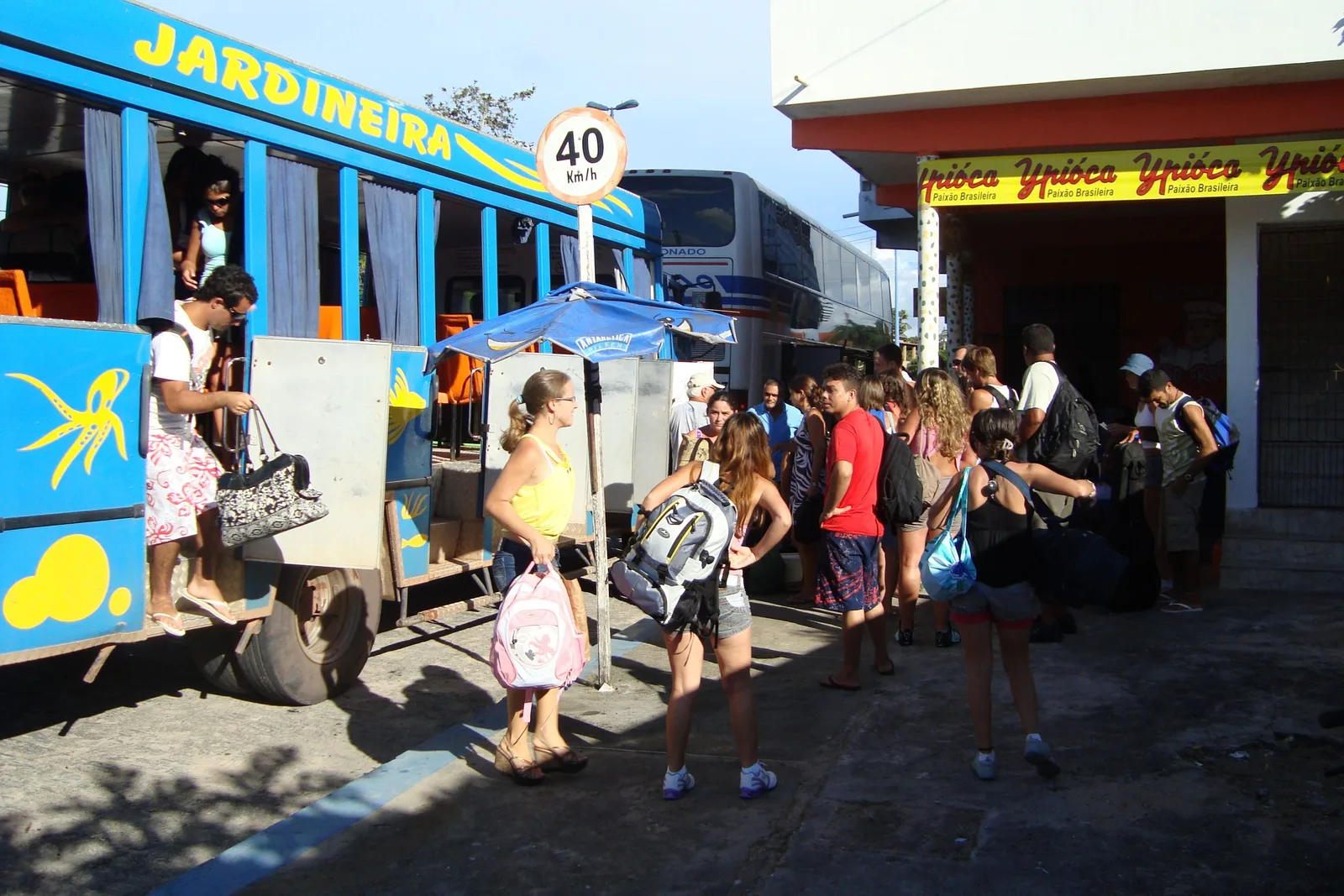 Passengers with bags boarding a blue open-sided 4x4 jardineira truck marked with its route, parked outside a shopfront in the sun