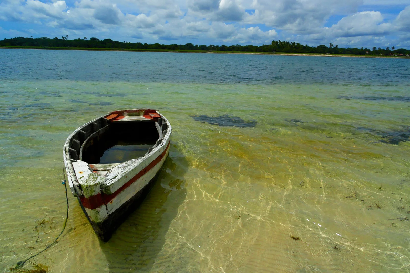 A small wooden boat moored in clear shallow lagoon water over pale sand, with low green vegetation along the far shore