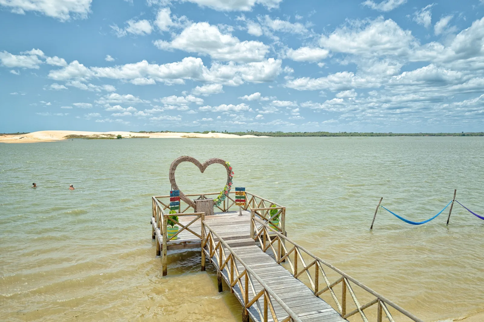 A wooden jetty running out into the shallow green Tatajuba lagoon, with a heart-shaped arch on it and hammocks strung over the water beside it
