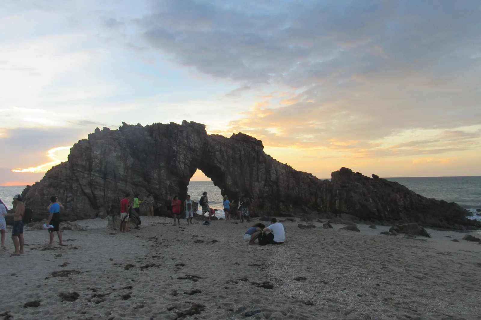 The Pedra Furada arch at the end of the day with people standing and sitting on the sand around it, the rock dark against a pale sky