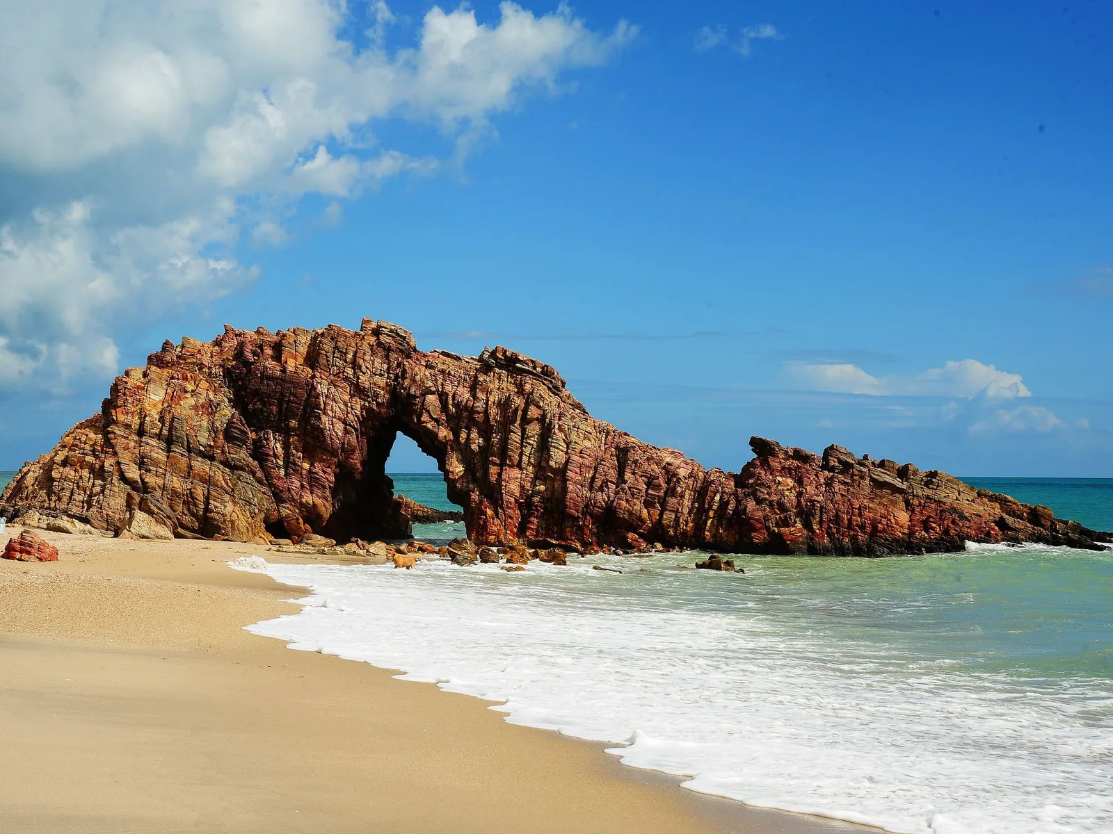 The Pedra Furada arch standing in shallow surf, banded orange and brown rock with a large opening through it, pale sand in front and turquoise water behind