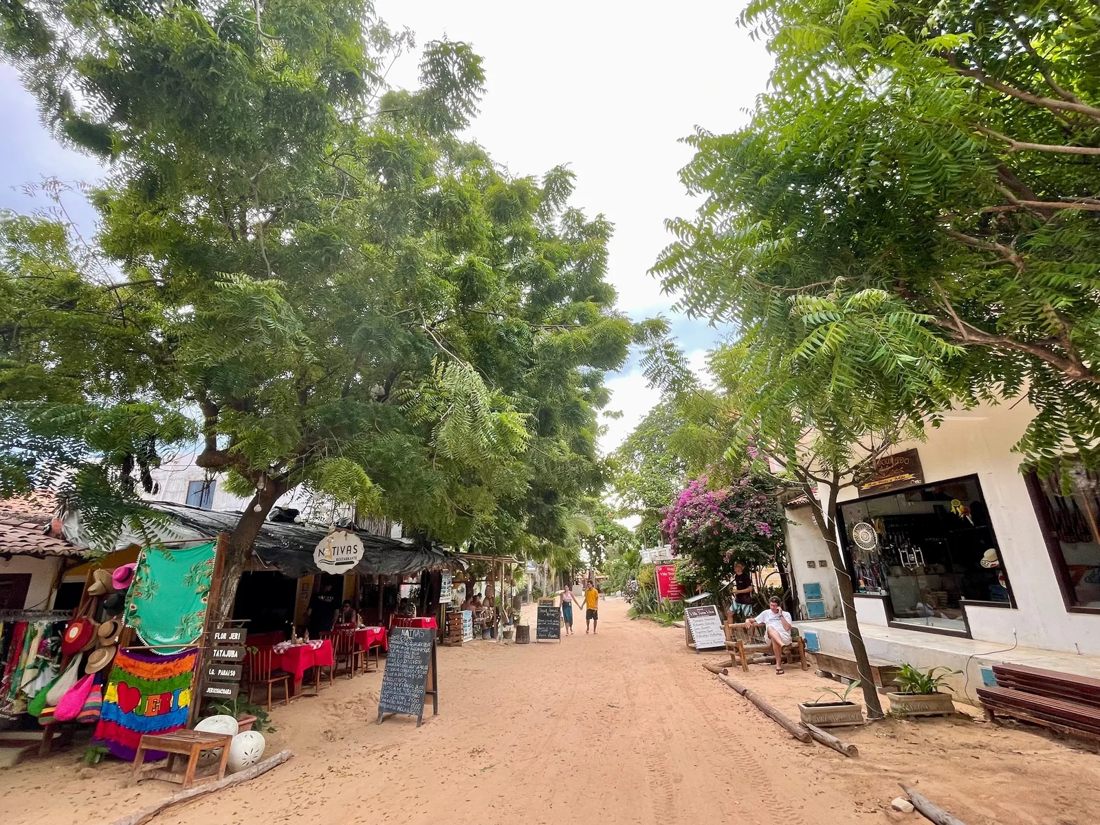A sand street in Jericoacoara lined with low shops and restaurants under leafy trees, with awnings, a rack of brightly colored hammocks and people walking in the middle of the street