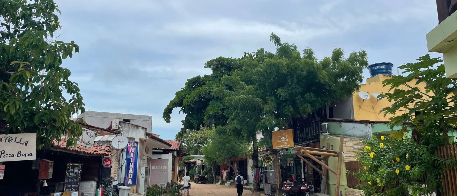 A sand street of low houses and small pousadas in Jericoacoara under leafy trees, with people walking along it and the sea visible at the far end