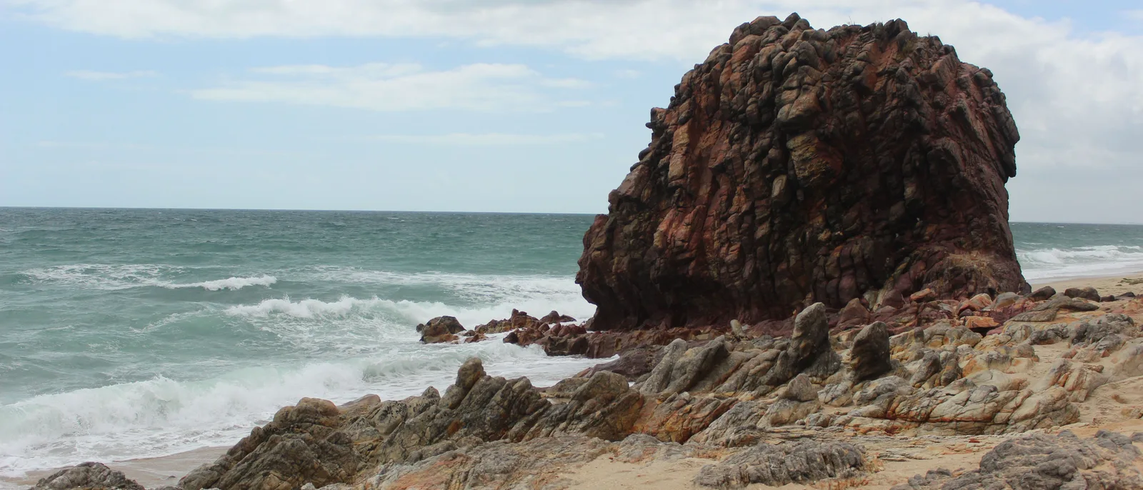 A wide view of the rocky coast at Praia da Malhada, dark stone outcrops standing in surf along a curve of pale sand with green scrub behind