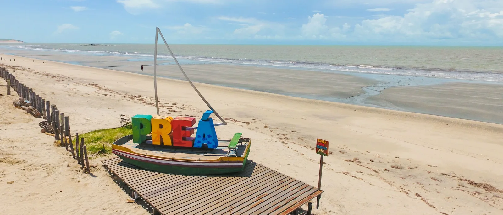 A large multicolored PREA letter sculpture standing on the sand at Praia do Prea, a small display boat propped beside it, a line of wooden posts along the dry sand to the left and a long empty shoreline running away behind