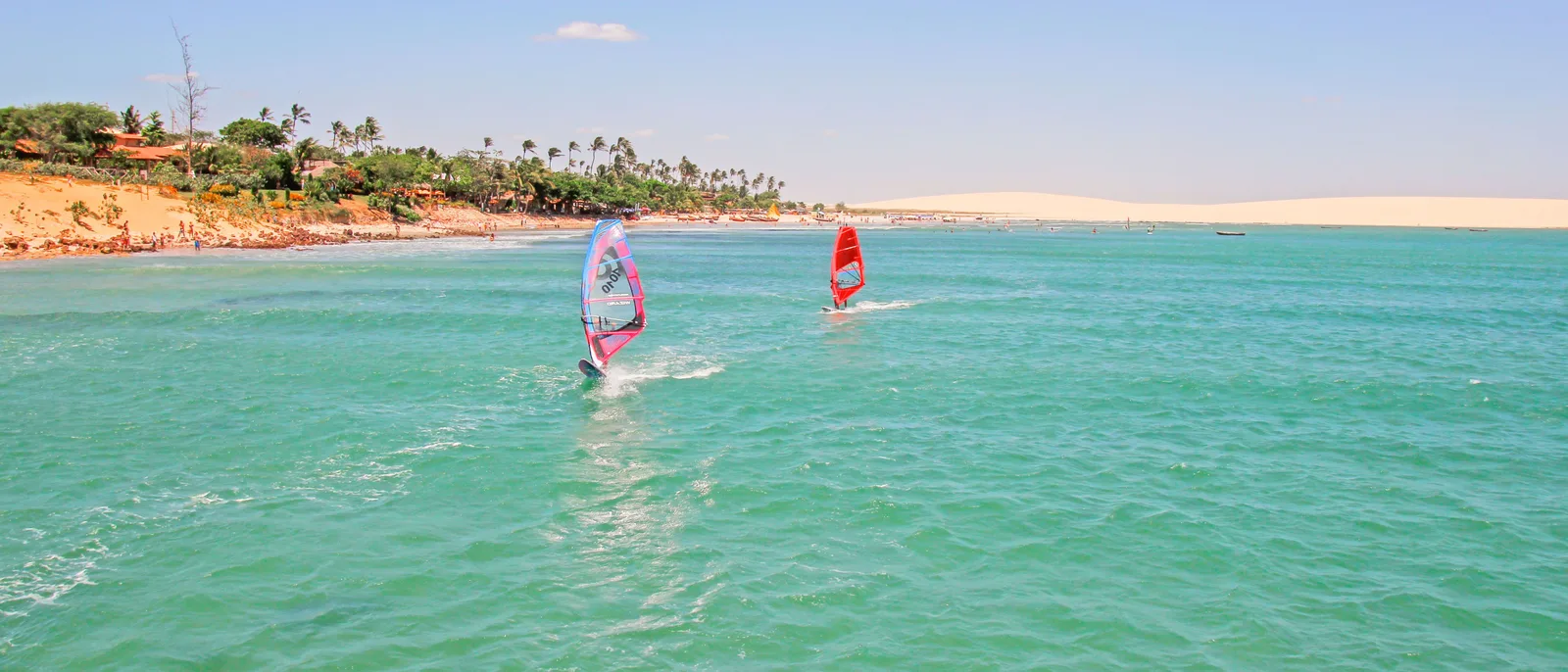 Two windsurfers on the clear turquoise water directly off the village, with the palm-lined shore and a pale dune ridge running away behind them
