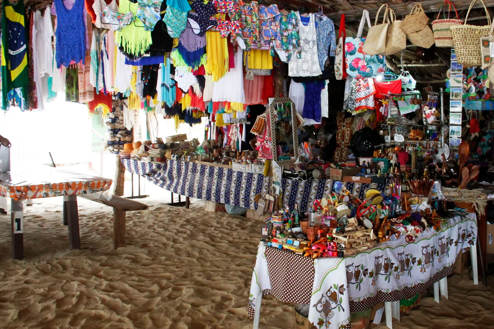 A craft stall set up on the sand under a canopy, hung with printed dresses, bags and towels, with tables of painted souvenirs and woven goods in front