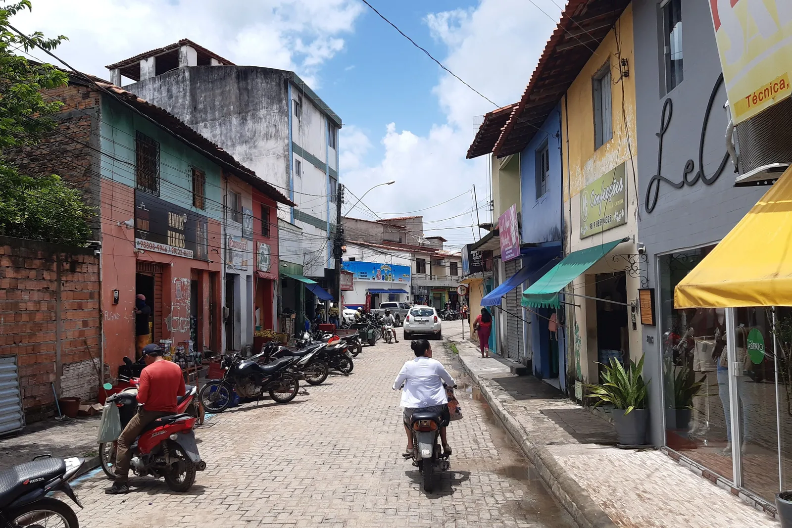 A street in central Barreirinhas lined with brightly painted shopfronts and awnings, motorbikes parked along both curbs and a rider coming up the middle