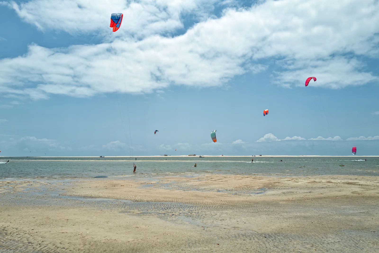 Kites in the air over shallow water and exposed sandbanks at the mouth of the Preguiças, riders spread out across a wide flat lagoon under a cloudy sky