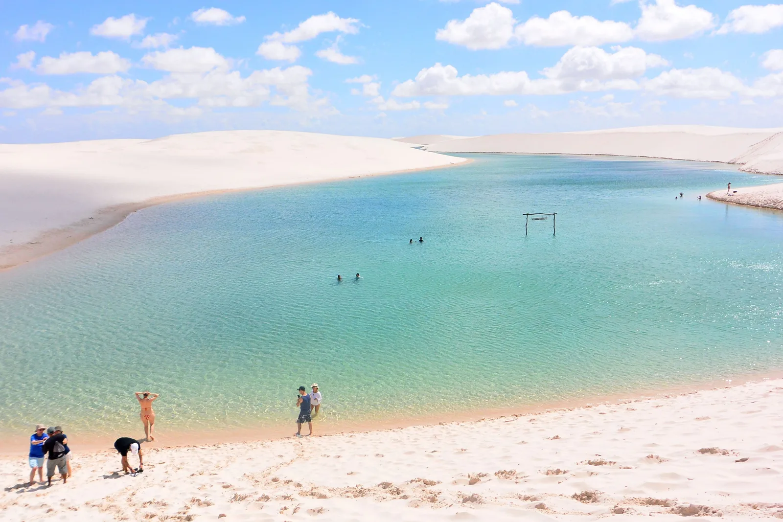 People standing and swimming in the clear turquoise shallows of Lagoa Azul, a broad sand beach in the foreground and white dunes closing the far side