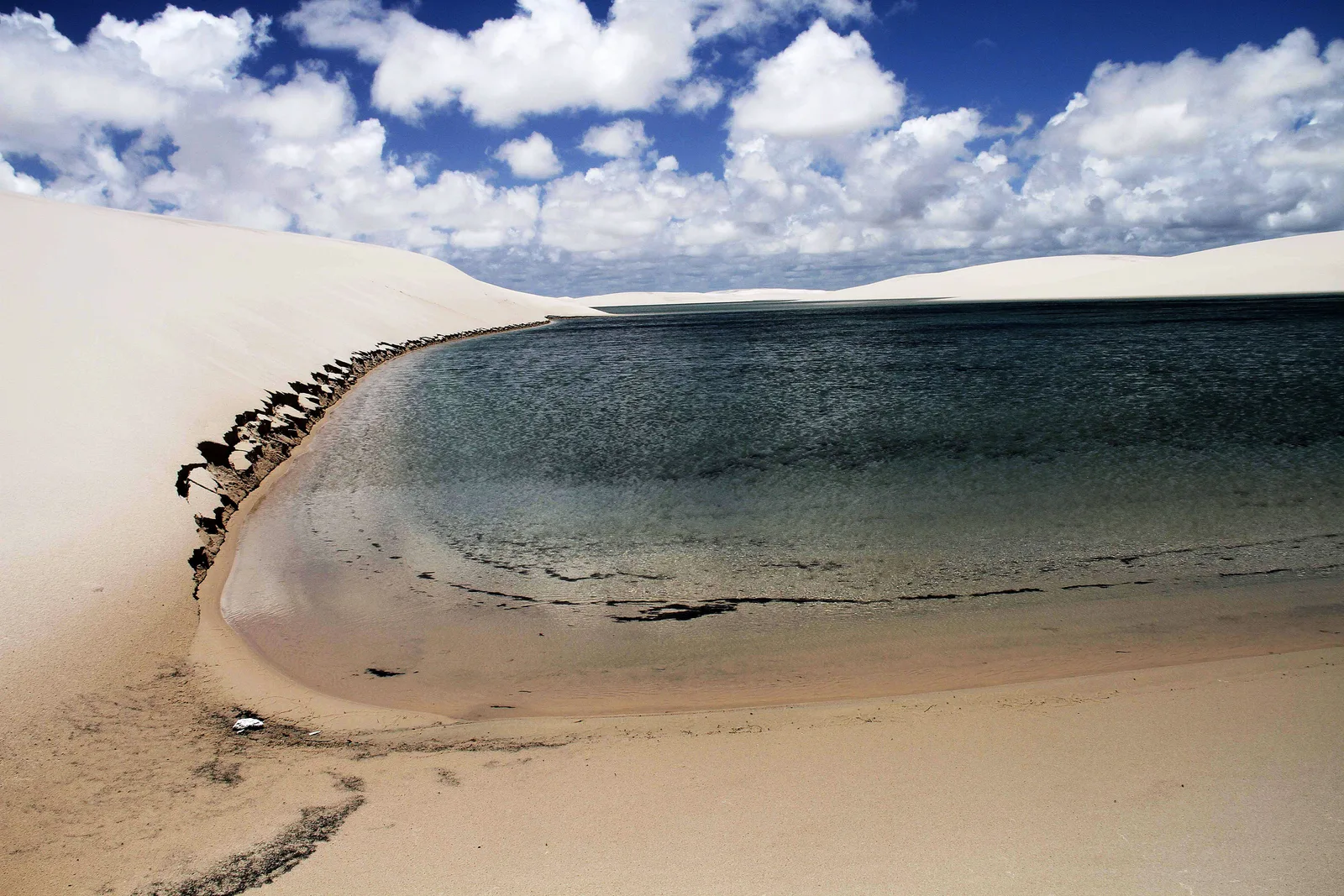 A lagoon at Santo Amaro with a dark ridge of stones running along its sandy edge, white dunes rising immediately behind the water