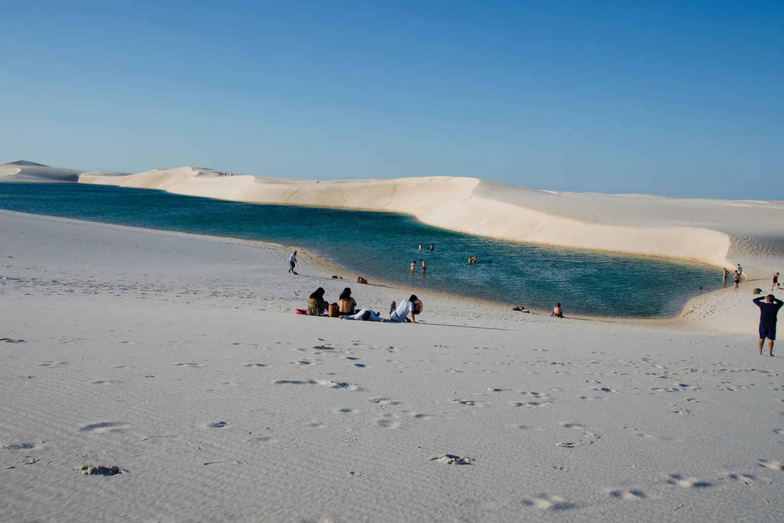 A turquoise lagoon set in blindingly white sand in the Pequenos Lençóis, with a handful of visitors standing at the water’s edge and dunes running away behind