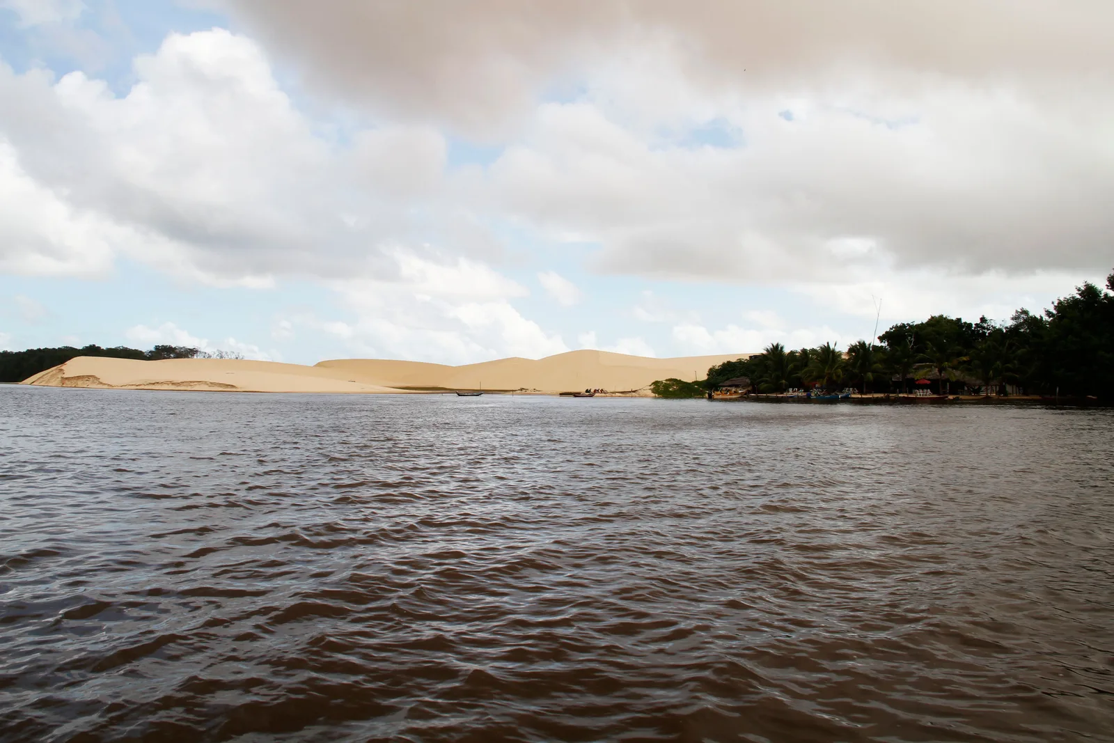 The Rio Preguiças seen from the water: brown river in the foreground, a green band of scrub along the bank and pale dunes rising behind it under a cloudy sky