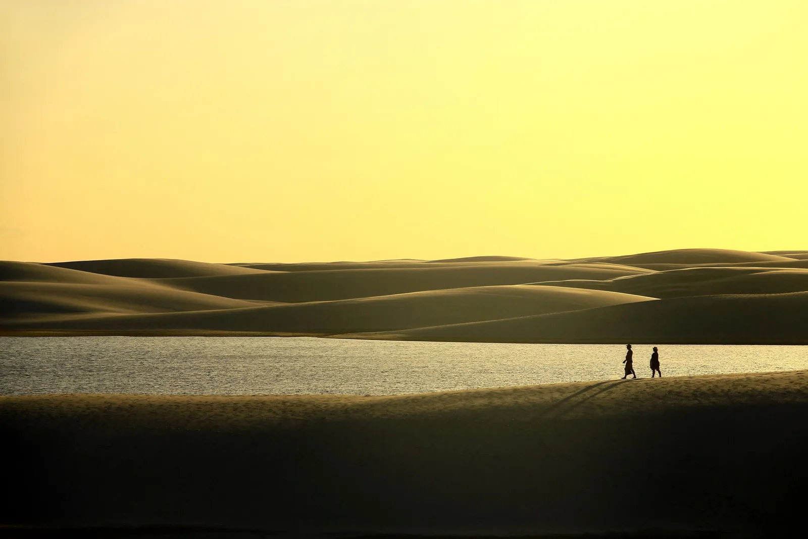 Two people in silhouette walking the sand at the edge of a lagoon against a sky burnt yellow by the setting sun, the dune field stacked behind them in bands of light and shadow