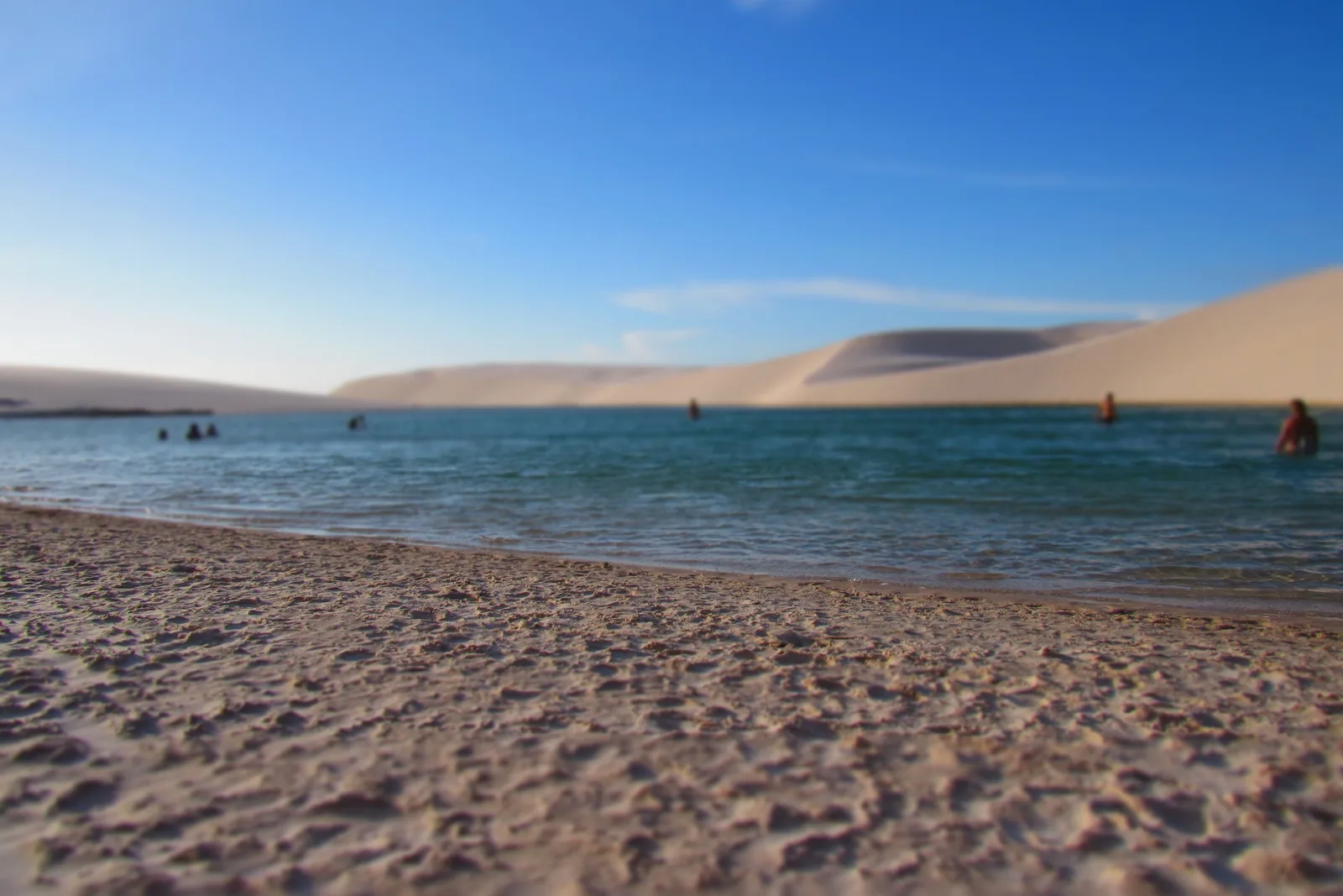 A wide beach of coarse pale sand running down to a big lagoon with swimmers standing in it, low white dunes along the whole far shore