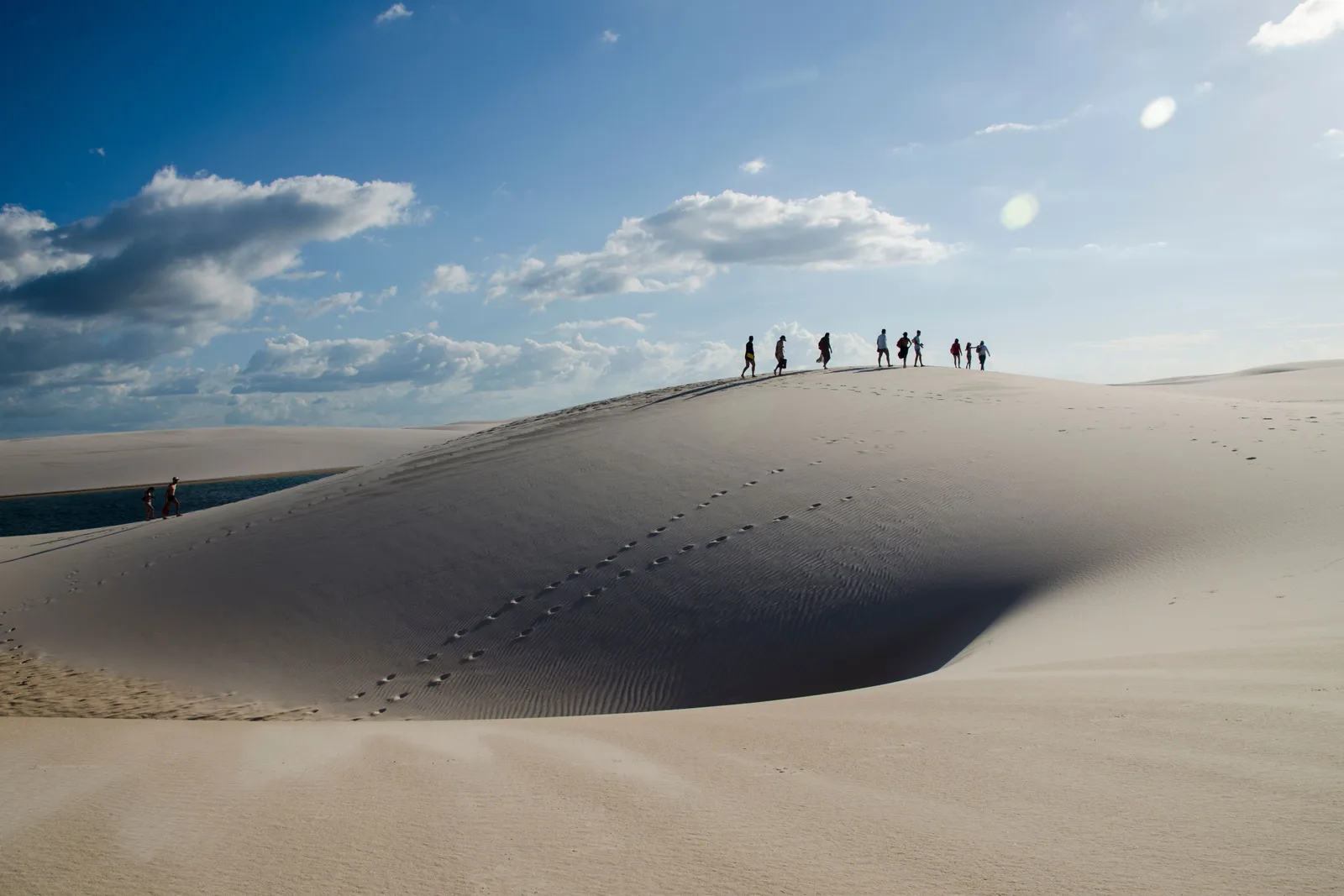 A line of people strung out along the crest of a white dune under a big sky of broken cloud, their footprints running back down the slope toward the camera